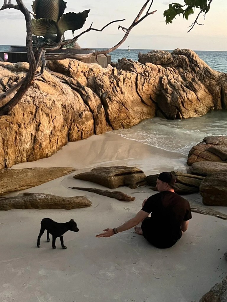 Ein junger Mann sitzt mit einem Hund am Strand am Meer auf Koh Samed