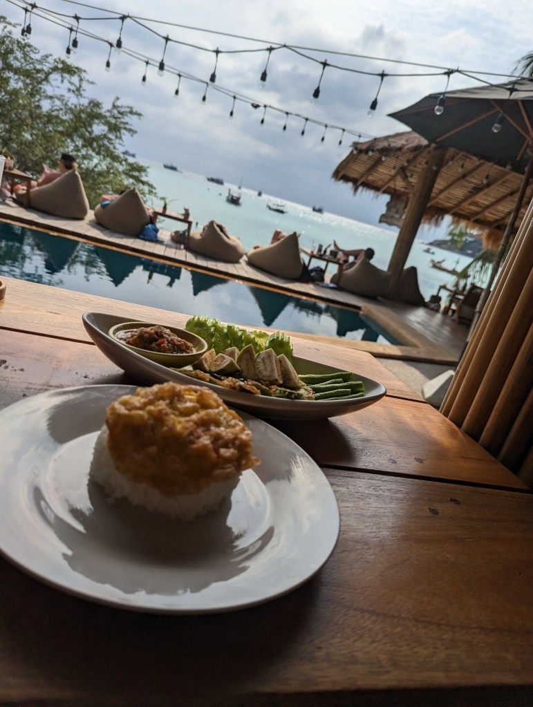 In einer thailändischen Beachbar auf Koh Tao sind auf einem Holztisch Kuchen und Snack. Dahinter ist das Meer mit Standhütten zu sehen.
