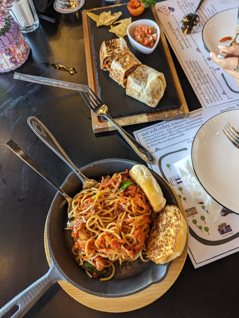 Thailändisches Nudelgericht steht in einer Pfanne auf einem Tisch. Daneben auf einem Teller Snacks.